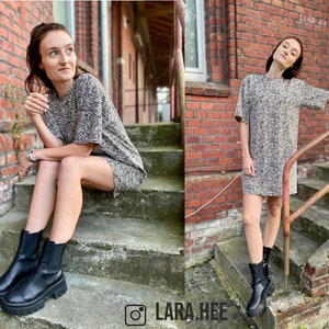 May include: A woman wearing a brown and white zebra print short-sleeved dress and black ankle boots. She is sitting on a set of stairs with a brick wall behind her.