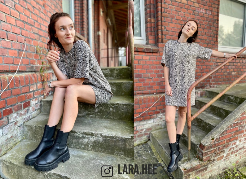 May include: A woman wearing a brown and white zebra print short-sleeved dress and black ankle boots. She is sitting on a set of stairs with a brick wall behind her.