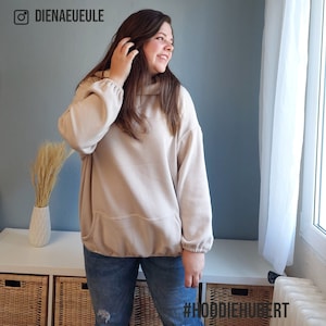 May include: A woman wearing a light beige oversized hoodie with a kangaroo pocket and a drawstring hood. She is wearing blue jeans and has long brown hair. The text #HOODIEMAKER is visible in the bottom right corner of the image.