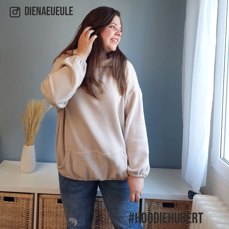 May include: A woman wearing a light beige oversized hoodie with a kangaroo pocket and a drawstring hood. She is wearing blue jeans and has long brown hair. The text #HOODIEMAKER is visible in the bottom right corner of the image.