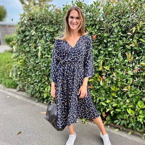 Puede incluir: Una mujer lleva un vestido midi estampado en negro y dorado con cuello en V y mangas 3/4. Lleva un bolso de mano de cuero negro y zapatillas blancas. El vestido tiene un cintur&oacute;n en la cintura. La foto fue tomada al aire libre frente a un seto verde. La leyenda en la parte inferior de la imagen dice "#SEWINGMACHINABERND".