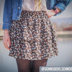 Pu&ograve; includere: Una donna indossa una giacca di jeans e una gonna a stampa floreale con un design a balze a strati. La gonna &egrave; nera con motivi floreali bianchi e blu. La donna indossa collant neri e una camicia bianca.