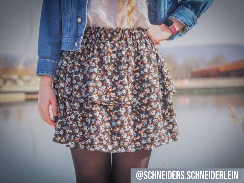 Pu&ograve; includere: Una donna indossa una giacca di jeans e una gonna a stampa floreale con un design a balze a strati. La gonna &egrave; nera con motivi floreali bianchi e blu. La donna indossa collant neri e una camicia bianca.