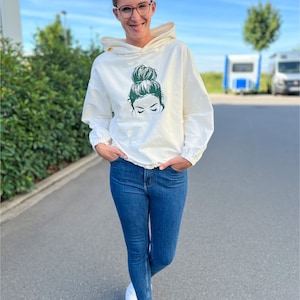 May include: A white hoodie featuring a green graphic of a woman with a bun on her head. The hoodie has a drawstring and is being worn by a person with blue jeans and white trainers. The text #HOODIEHUBERT is visible at the bottom of the image.