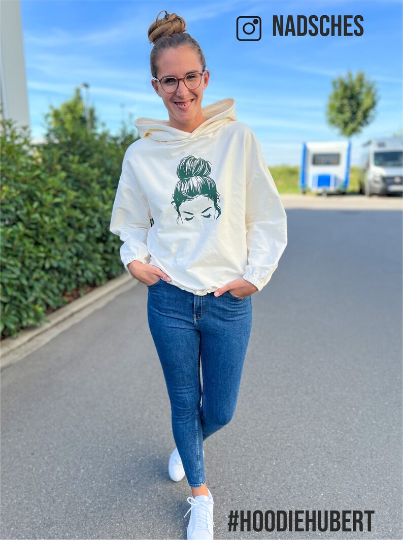 May include: A white hoodie featuring a green graphic of a woman with a bun on her head. The hoodie has a drawstring and is being worn by a person with blue jeans and white trainers. The text #HOODIEHUBERT is visible at the bottom of the image.