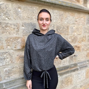 May include: A woman wearing a grey and black leopard print hoodie with a drawstring tie. The hoodie has long sleeves and a relaxed fit. She is also wearing black leggings. The image includes the hashtag #HOODIEHUBERT.