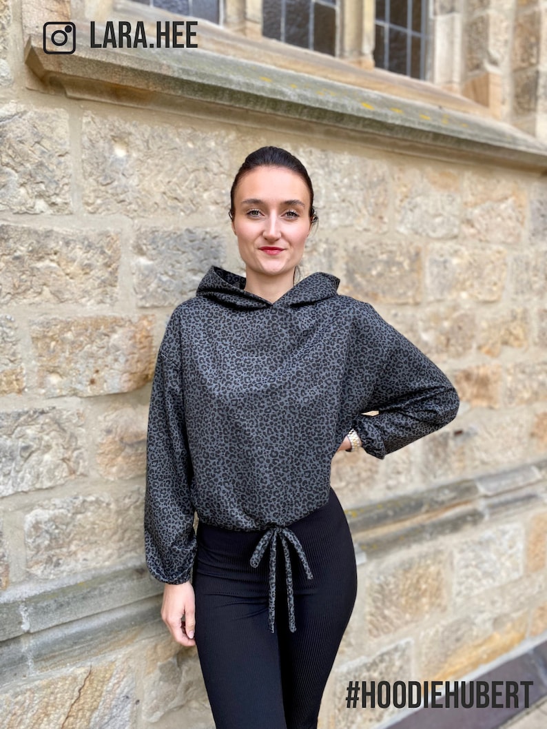May include: A woman wearing a grey and black leopard print hoodie with a drawstring tie. The hoodie has long sleeves and a relaxed fit. She is also wearing black leggings. The image includes the hashtag #HOODIEHUBERT.