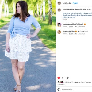 Pu&ograve; includere: Una donna indossa una blusa azzurra annodata in vita e una gonna bianca a strati con un motivo floreale. &Egrave; in piedi su un sentiero lastricato con alberi verdi su entrambi i lati. L'immagine proviene da un post sui social media con il testo "smalino.de Und nochmal in voller Pracht #werbung #n&auml;hen #smalino #derponyhof #lochstrick #melange #Designn&auml;hen #wraptopwalter #miniskirtmanfred #Sewingmachina 151 Wo"