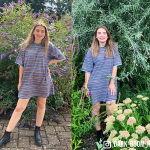 May include: A woman wearing a striped blue and multi-colored t-shirt dress with black boots. She is standing in front of a green bush with purple flowers.