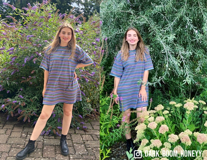 May include: A woman wearing a striped blue and multi-colored t-shirt dress with black boots. She is standing in front of a green bush with purple flowers.