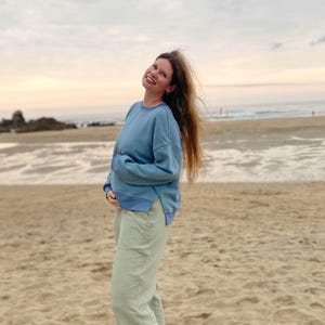 May include: A woman wearing a light blue sweatshirt and light green pants stands on a sandy beach. The sky is a soft orange and pink. The text "KURZ/CROPPED #SEWINGMACHINAANTON" is at the bottom of the image.
