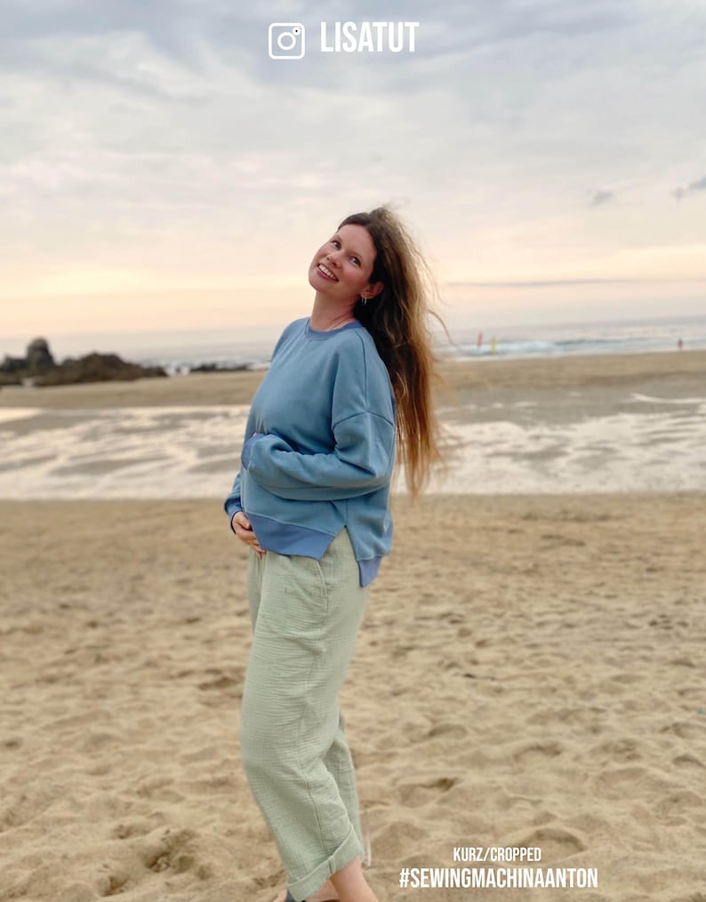 May include: A woman wearing a light blue sweatshirt and light green pants stands on a sandy beach. The sky is a soft orange and pink. The text "KURZ/CROPPED #SEWINGMACHINAANTON" is at the bottom of the image.
