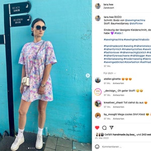 May include: A woman wearing a tie-dye dress with a white purse and white sneakers. The dress has a loose fit and is made of a soft, comfortable fabric. The background is a blue wall with signs for a museum, beach, and station.