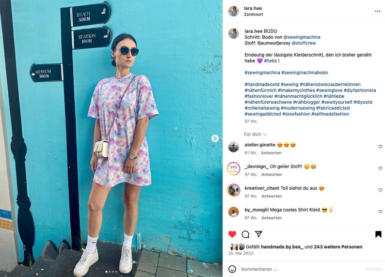 May include: A woman wearing a tie-dye dress with a white purse and white sneakers. The dress has a loose fit and is made of a soft, comfortable fabric. The background is a blue wall with signs for a museum, beach, and station.
