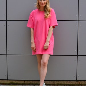 May include: A woman wearing a pink oversized t-shirt dress and white sneakers. She is standing in front of a gray wall.