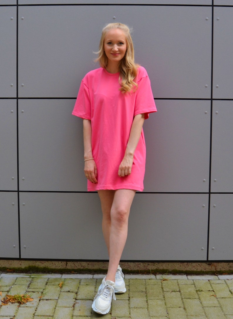 May include: A woman wearing a pink oversized t-shirt dress and white sneakers. She is standing in front of a gray wall.