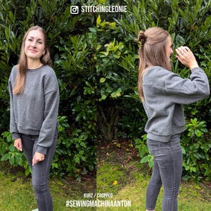 May include: A gray cropped sweatshirt with long sleeves and a ribbed hem. The sweatshirt is being worn by a person with long brown hair. The person is standing in front of a green leafy background.