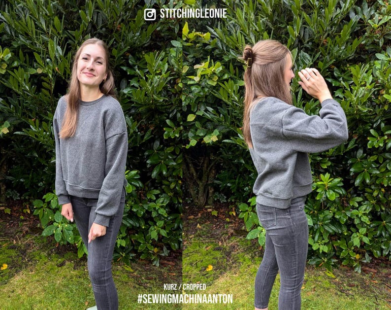 May include: A gray cropped sweatshirt with long sleeves and a ribbed hem. The sweatshirt is being worn by a person with long brown hair. The person is standing in front of a green leafy background.