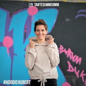 May include: A woman wearing a light beige hoodie with a drawstring at the bottom. The hoodie has a relaxed fit and a slightly cropped length. The woman is standing in front of a graffiti wall with the text "#HOODIEHUBERT" and "TANTESUMMSUMM" visible.