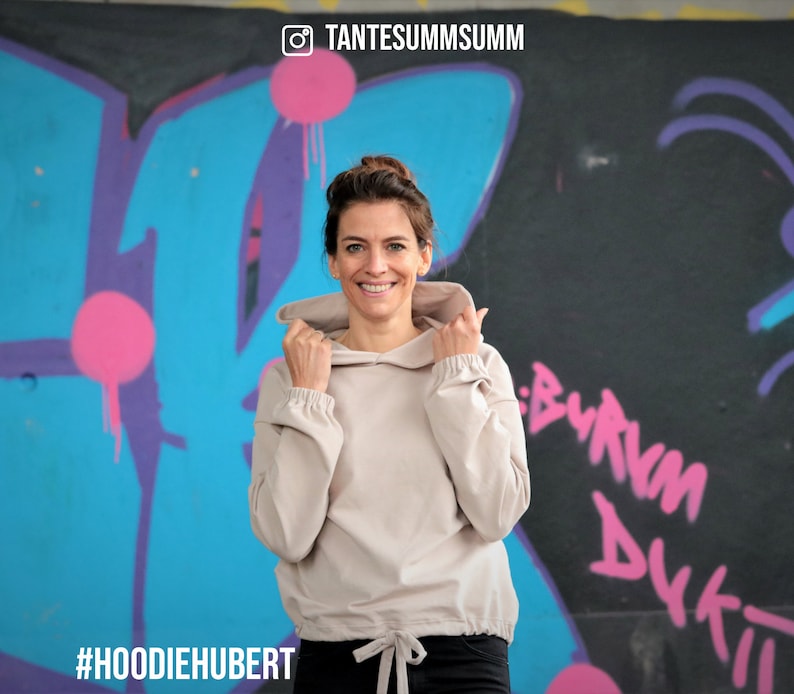 May include: A woman wearing a light beige hoodie with a drawstring at the bottom. The hoodie has a relaxed fit and a slightly cropped length. The woman is standing in front of a graffiti wall with the text "#HOODIEHUBERT" and "TANTESUMMSUMM" visible.