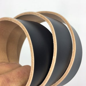 May include: Close-up of a rolled black leather belt with a natural tan edge. The leather is smooth and the edges are neatly finished. The belt is likely for use with trousers or skirts.