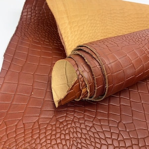Könnte beinhalten: Eine Rolle braunes Leder mit Krokodilhautstruktur. Das Leder hat einen satten, warmen Braunton, mit einer helleren, beigen Farbe auf der Unterseite. Das Leder ist teilweise aufgerollt und zeigt die Textur und Farbe.