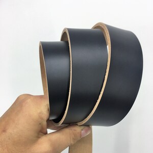May include: A rolled black leather belt with a natural tan underside. The belt is held in a hand against a white background, highlighting the smooth texture and colour contrast.