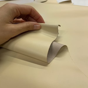 May include: A close-up of a piece of off-white leather being folded and held by a hand. The leather is smooth and has a slight sheen.