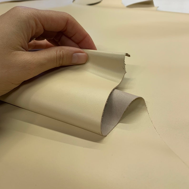 May include: A close-up of a piece of off-white leather being folded and held by a hand. The leather is smooth and has a slight sheen.