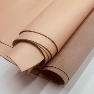 Puede incluir: Dos rollos de cuero, uno en color beige claro y el otro en textura de ante marrón medio. El cuero beige está enrollado apretado, revelando los bordes del material. El cuero de ante está enrollado suelto.