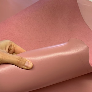 May include: A close-up of a soft, pink leather fabric. The fabric is smooth and has a slight sheen. It is being held in a hand.