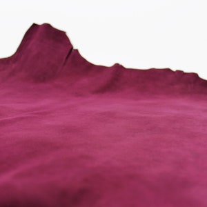 May include: A piece of burgundy-colored leather with a soft, suede-like texture. The edges are uneven, suggesting a natural cut. The material is displayed against a plain white background, highlighting its rich color and texture.