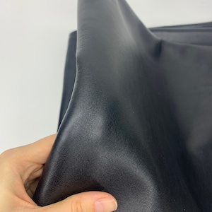 May include: A piece of black faux leather fabric with a smooth, slightly grained texture. The material is flexible and pliable, showing soft folds and creases across its surface. The dark, matte finish reflects light subtly along the curved edges.