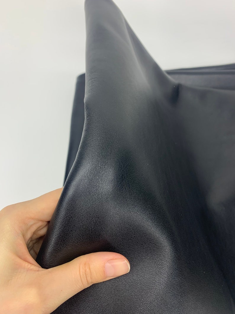 May include: A piece of black faux leather fabric with a smooth, slightly grained texture. The material is flexible and pliable, showing soft folds and creases across its surface. The dark, matte finish reflects light subtly along the curved edges.