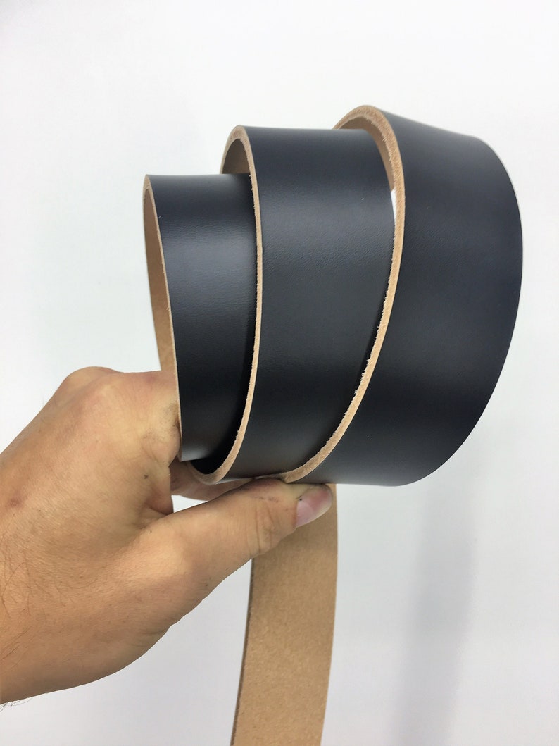 May include: A rolled black leather belt with a natural tan underside. The belt is held in a hand against a white background, highlighting the smooth texture and colour contrast.