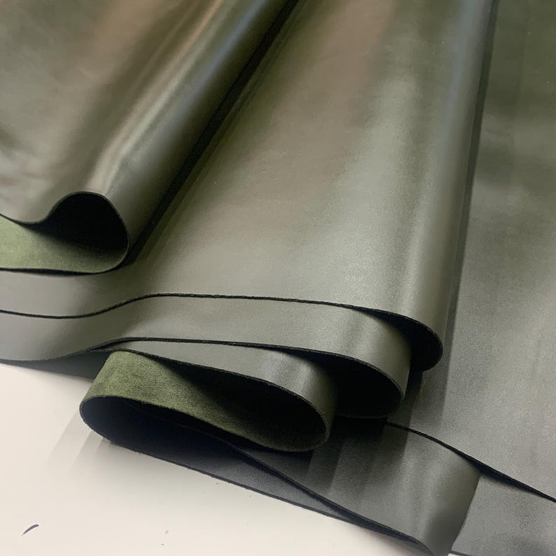 May include: Close-up of olive green leather fabric, rolled and layered. The leather has a smooth, matte finish with subtle variations in color and texture. The underside of the leather is a darker green suede-like material.