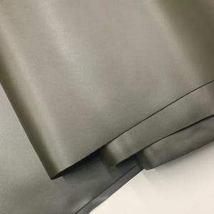 May include: Close-up of olive green leather material. The leather has a smooth, matte finish and appears to be folded over itself, revealing the underside. The color is a muted, earthy tone, suitable for various crafting projects.