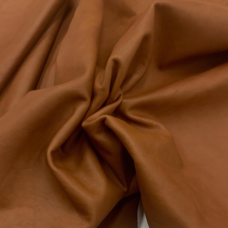 Puede incluir: Primer plano de un trozo de cuero marr&oacute;n con una textura suave y lisa. El cuero est&aacute; doblado y drapeado, mostrando su flexibilidad.