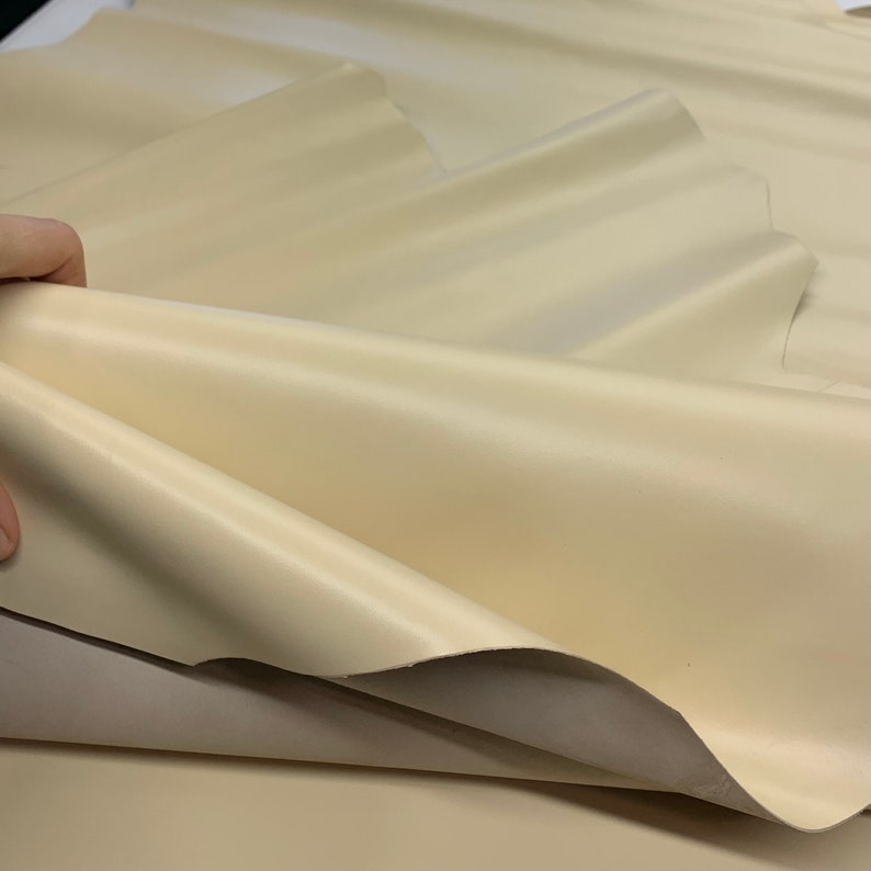 May include: Cream coloured leather fabric folded in a soft, wavy pattern. The fabric is smooth and has a subtle sheen.