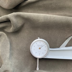 Può includere: Primo piano di un campione di tessuto scamosciato verde oliva. Un calibro a quadrante argentato, contrassegnato "CALATI MADE IN ITALY", misura lo spessore del tessuto. Il calibro indica circa 0,1 mm.