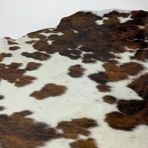 Pode incluir: Close-up de um tapete de pele de vaca com um padrão natural. O tapete apresenta uma mistura de manchas castanhas e brancas, criando uma estética única e rústica. A textura da pele é visível, adicionando ao seu apelo orgânico. Ideal para decoração de casa.