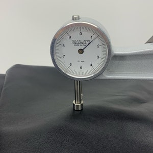 May include: A silver Calati thickness gauge with a white circular dial face, marked 0.1 mm, measuring the thickness of a smooth, matte black leather material. The gauge features a needle pointing near the 1 mark on the scale.