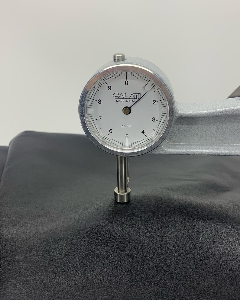May include: A silver Calati thickness gauge with a white circular dial face, marked 0.1 mm, measuring the thickness of a smooth, matte black leather material. The gauge features a needle pointing near the 1 mark on the scale.