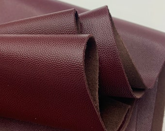 Op de afbeelding: Close-up van gevouwen bordeauxrood leer met een getextureerd, korrelig oppervlak. Het leer is gelaagd en onthult een donkerbruine onderkant. De belichting benadrukt de textuur en kleurvariaties van het materiaal.