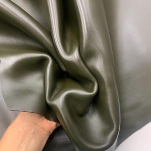 May include: Close-up of olive green leather fabric with a smooth, shiny surface. The leather is folded, showing the texture and sheen. The color is a deep, muted green, with subtle variations in tone.