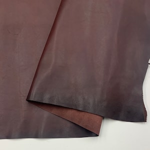 Könnte beinhalten: Ein Stück dunkelbraunes Leder mit einer glatten, leicht strukturierten Oberfläche. Das Leder ist in der Mitte gefaltet und zeigt eine hellere braune Unterseite.