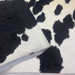 May include: A close-up of a cowhide rug with a black and white spotted pattern. The rug is folded over, revealing the underside. The hide has a soft, textured appearance.