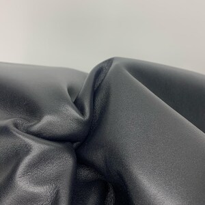 May include: A close-up view of a piece of smooth, matte black leather material. The surface is folded and gathered, creating deep, soft curves and shadows that highlight the texture and flexibility of the dark, solid-coloured material.