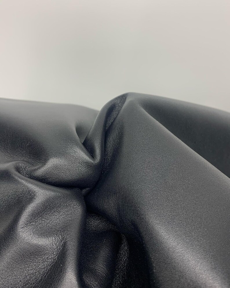 May include: A close-up view of a piece of smooth, matte black leather material. The surface is folded and gathered, creating deep, soft curves and shadows that highlight the texture and flexibility of the dark, solid-coloured material.