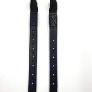 Può includere: Due cinturini in pelle nera con fori numerati per regolare la lunghezza. Ogni cinturino ha un anello nero in alto.